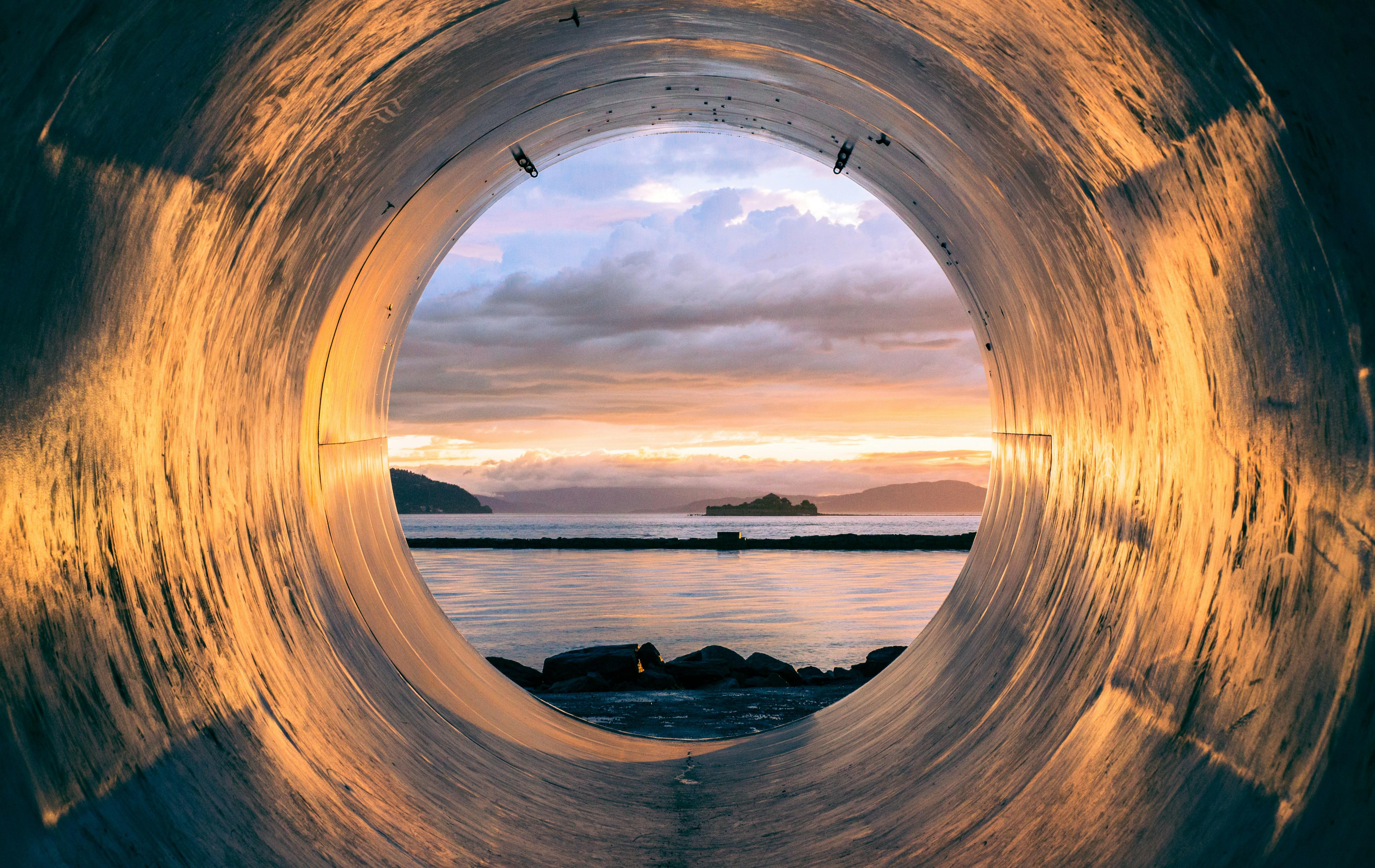 View through a large industrial pipe looking out at a body of water during sunset